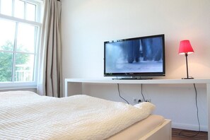 A bedroom with a bed, a television mounted on a white wall, and a red lamp. - Holiday apartment with 2 bedrooms (Norderney)