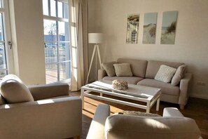 A living room with a sofa, armchairs, a coffee table, and a lamp. There are three paintings on the wall. - Holiday apartment with 2 bedrooms (Norderney)