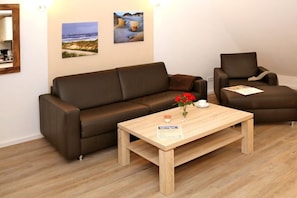 A modern living room with a brown leather sofa, a wooden coffee table with a vase of red flowers, and two framed landscape paintings on the wall. - House Hahnen - Upper Floor (Norderney)