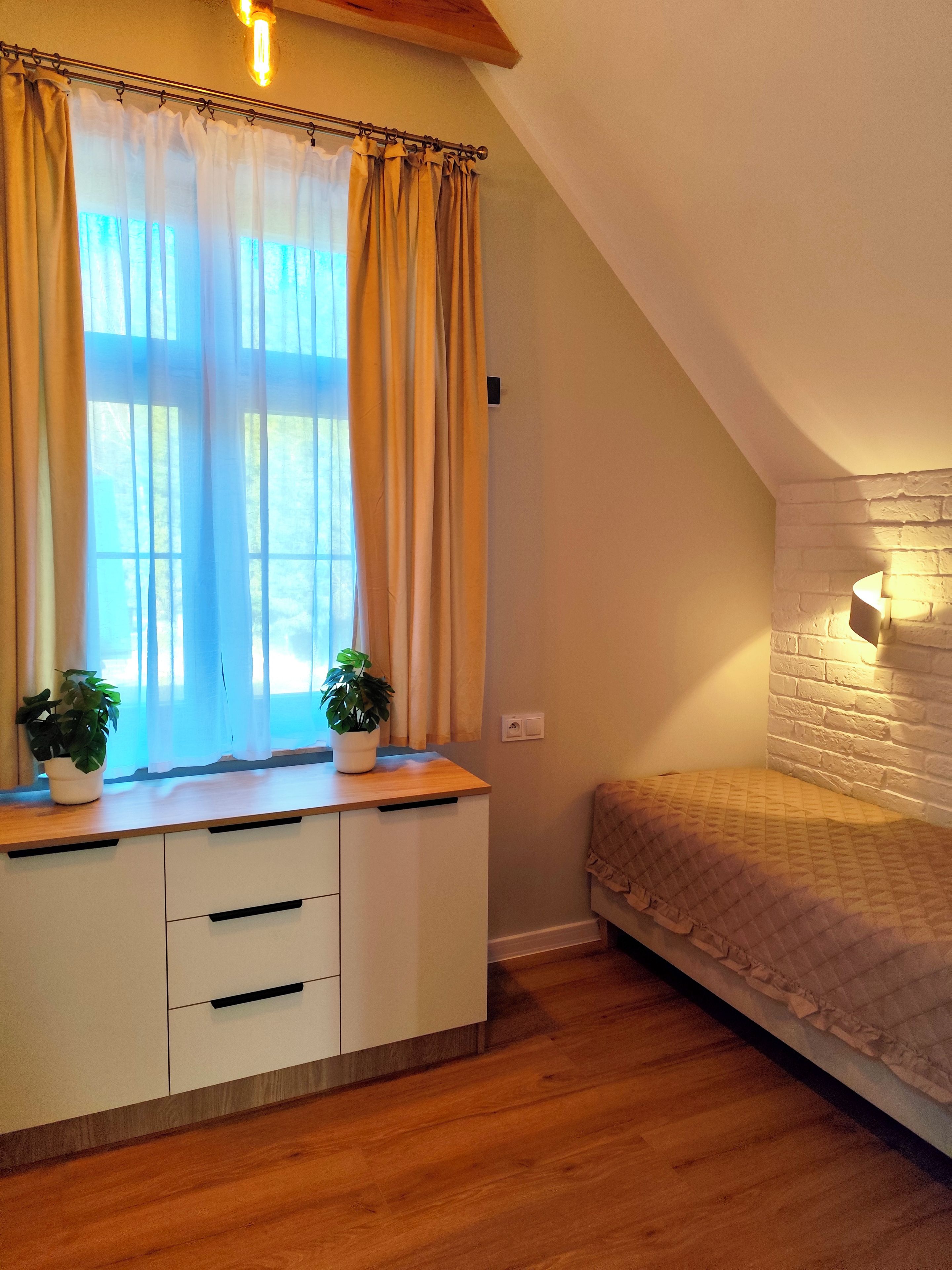 A bedroom with a wooden bed, a white dresser with drawers, a window with curtains, and a brick wall.
