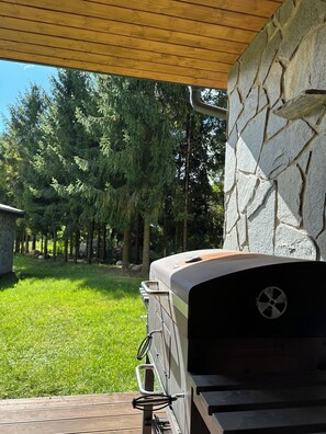 Un barbecue avec un mur de pierres en arrière-plan, une terrasse en bois et une cour verdoyante avec des arbres.