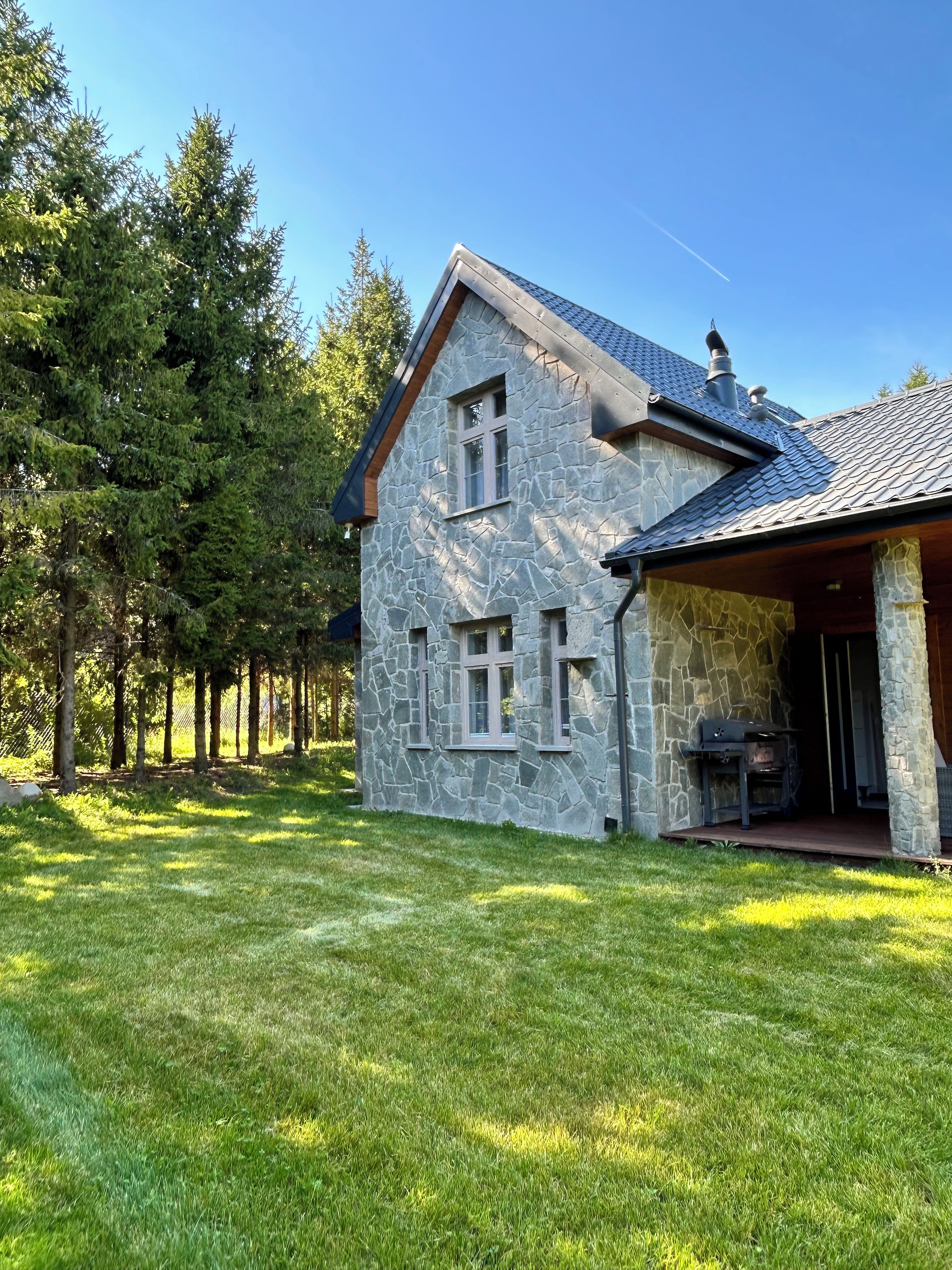 Une maison en pierre dotée d’un porche abrité et d ’un barbecue, entourée d’une pelouse bien entretenue et de grands arbres.