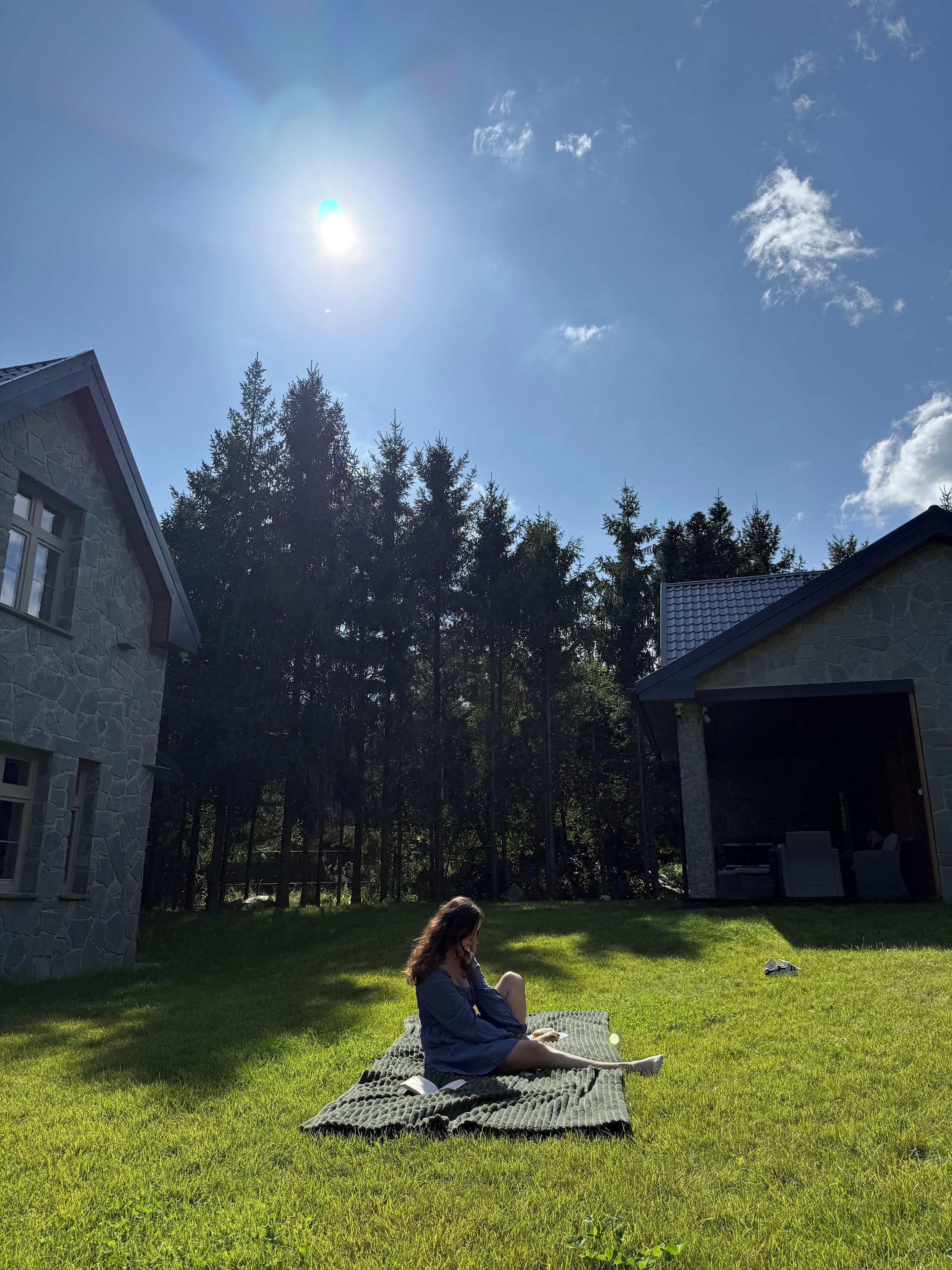 A person sitting on a blanket in a grassy area with a house and trees in the background.