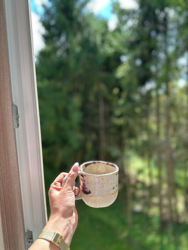 Une main tenant une tasse près d’une fenêtre donnant sur une forêt.
