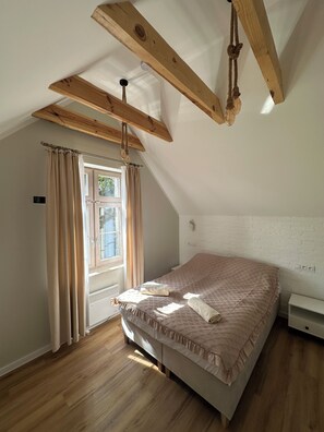 Une chambre avec un plafond en pente, des poutres en bois, un lit recouvert d’une courtepointe matelassée, un radiateur et une fenêtre avec des rideaux.