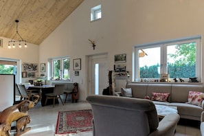 A spacious living room with a high ceiling, a dining area with a table and chairs, and large windows offering a view of greenery outside. - Holiday home in Bell with swimming pool (Bell)