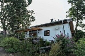 Una casa con fachada blanca, un porche de madera y un jardín con diversas plantas y una estatua.