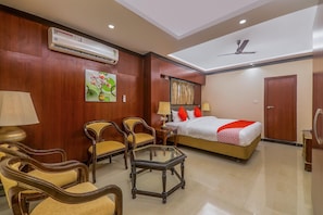 Habitación de hotel con cama, dos sillas, una mesa, equipo de aire acondicionado y ventilador de techo.