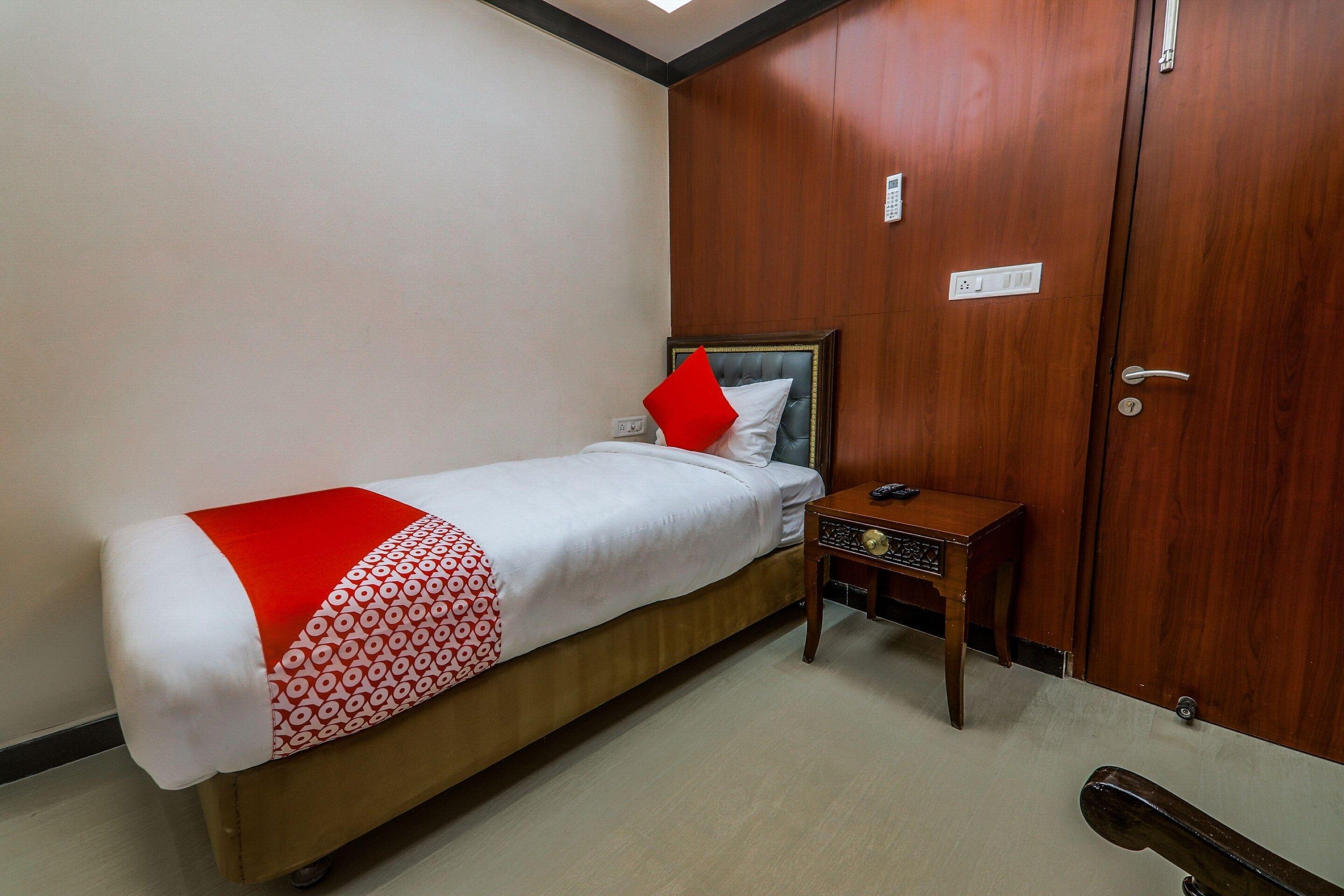 Una cama individual con un almohadón rojo, una mesita de noche con un control remoto y un armario de madera con un tirador.