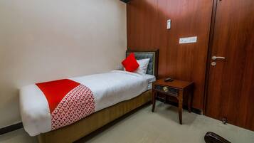 Una cama individual con un almohadón rojo, una mesita de noche con un control remoto y un armario de madera con un tirador.