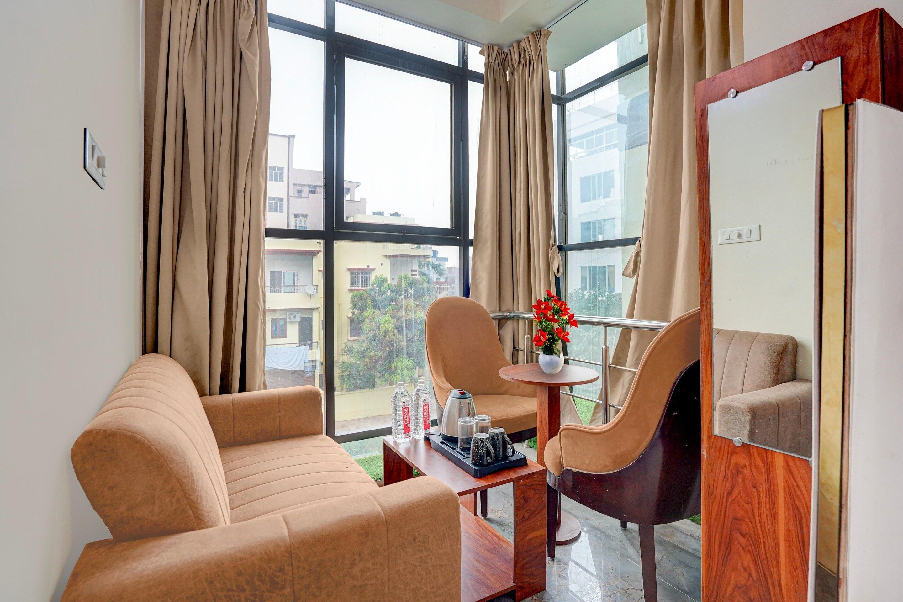 A hotel room with a sofa, armchairs, a small table with a vase of flowers, and a view of buildings through large windows.