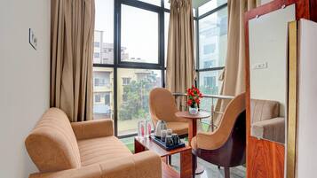 A hotel room with a sofa, armchairs, a small table with a vase of flowers, and a view of buildings through large windows.