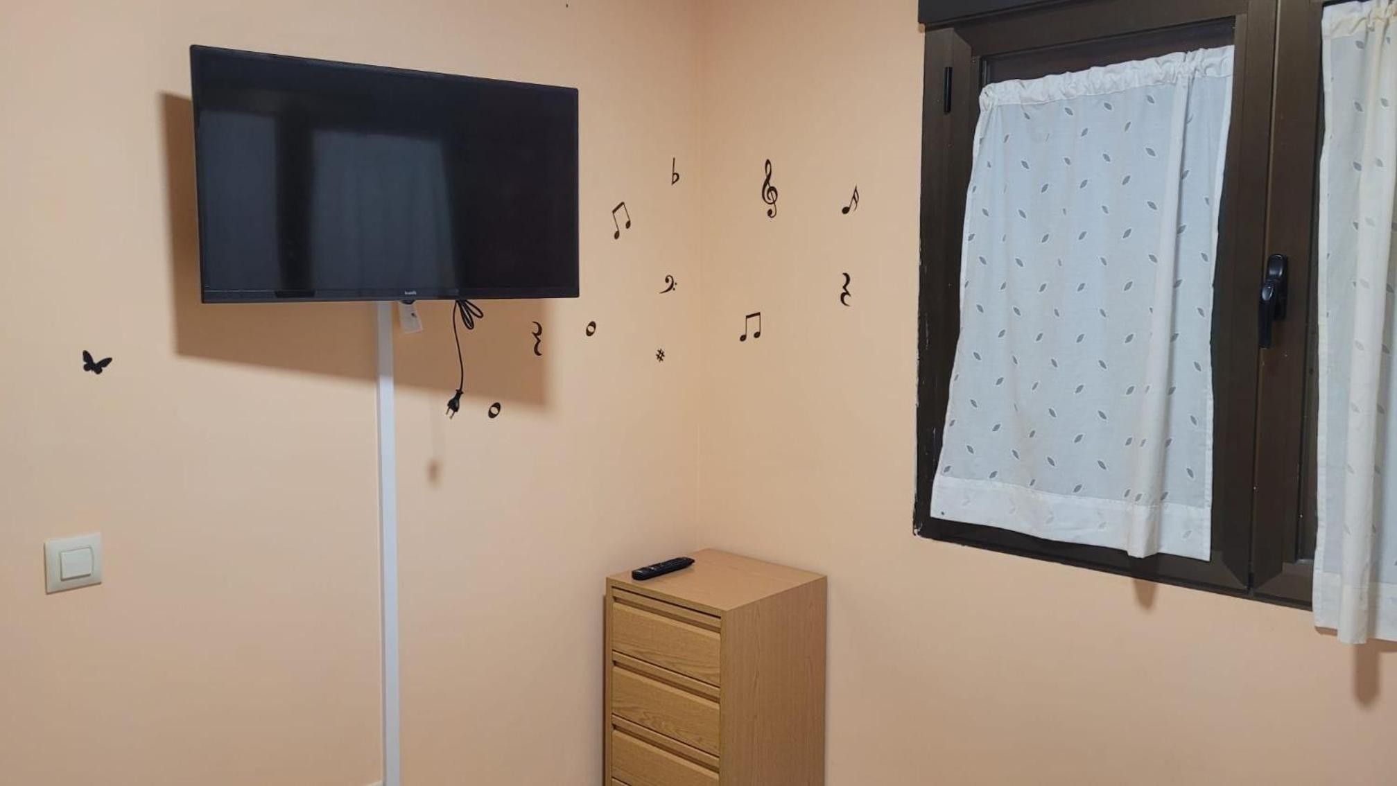 Une pièce avec un téléviseur fixé au mur, une fenêtre avec un rideau et un meuble en bois.
