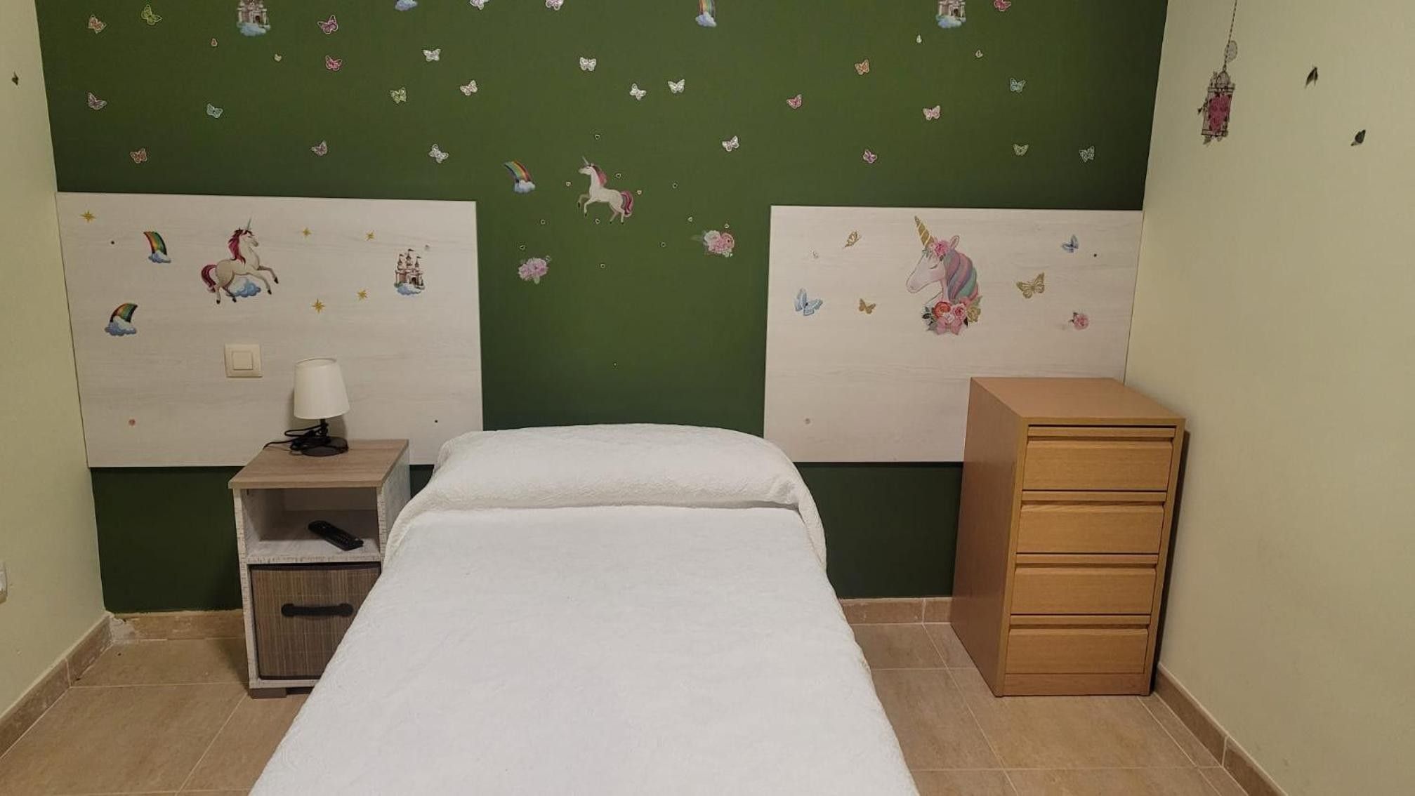 Un dormitorio con una cama, una mesita de noche con lámpara, un aparador y adhesivos con forma de mariposas en la pared.