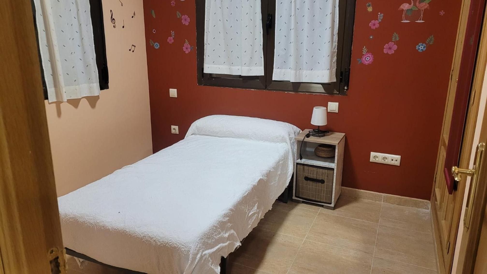 Una habitación pequeña y bien arreglada con una cama individual, una mesita de noche con lámpara y una ventana con cortinas.