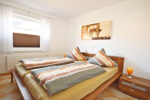 A bedroom with two beds, a bedside table with a lamp, and a window with blinds. - Holiday apartment with 1 bedroom (Norddeich)