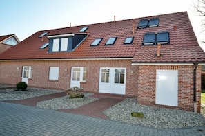A two-story brick building with a sloped roof, multiple windows, and a paved driveway. - Holiday apartment with 1 bedroom (Norddeich)