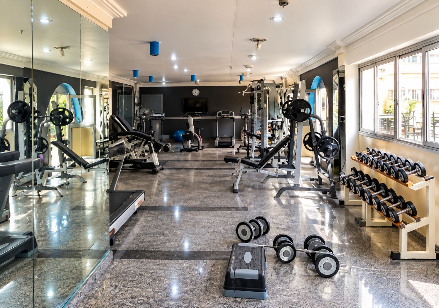 Un gimnasio con varios equipos de ejercicio, un muro de espejos y una ventana que proporciona luz natural.