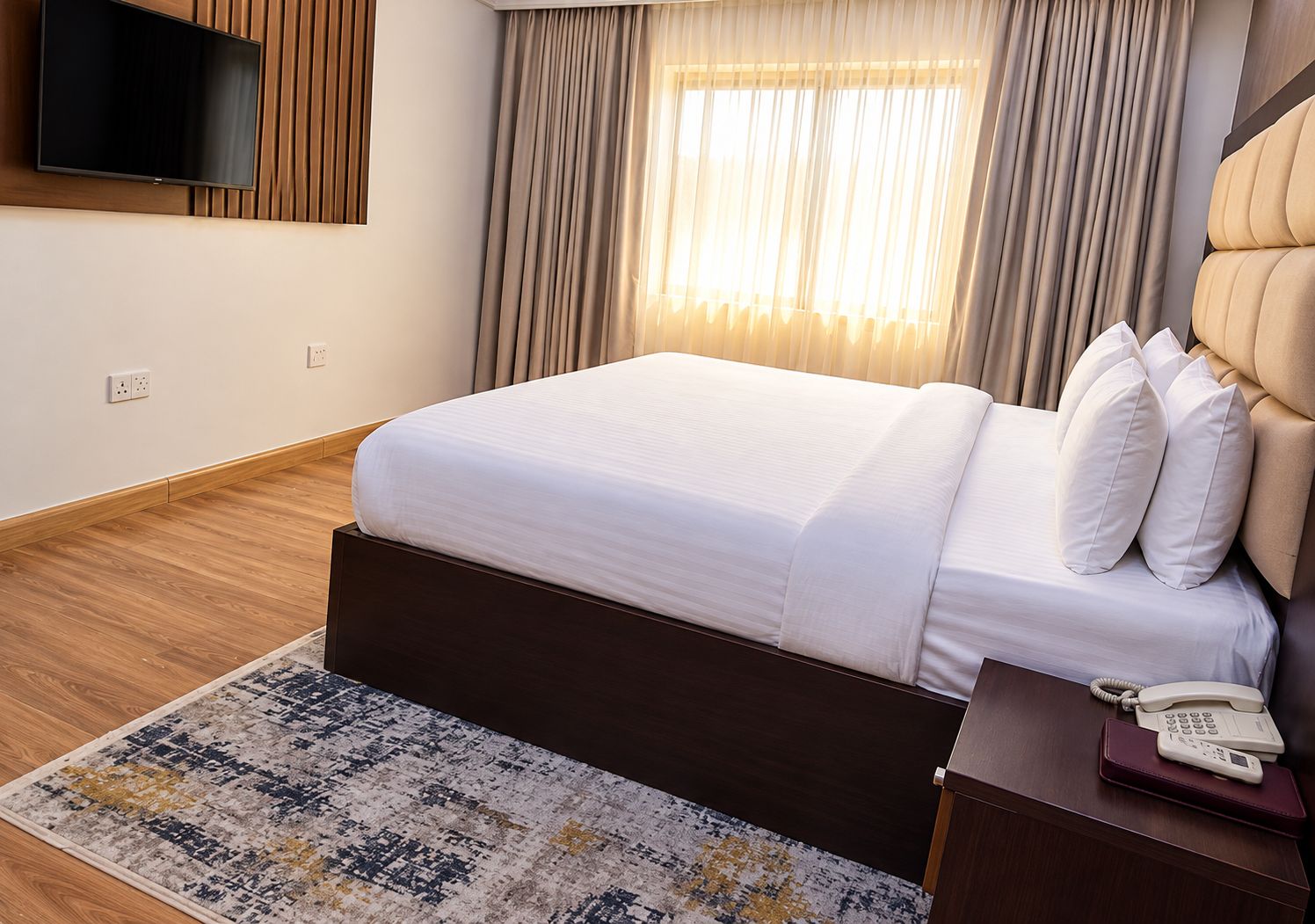 Quarto de hotel com cama, televisão na parede, mesa de cabeceira com telefone e tapete estampado.