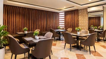 Uma sala de conferências de hotel com várias mesas e cadeiras, uma janela grande com persianas e um aparelho de ar condicionado fixado na parede.