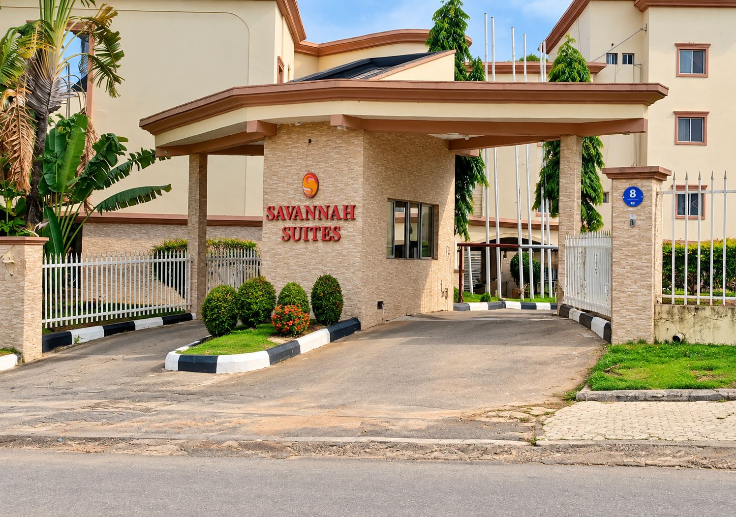 Entrada de hotel com um letreiro indicando 'Savannah Suites', uma entrada com portão e área ajardinada com arbustos pequenos e uma palmeira.