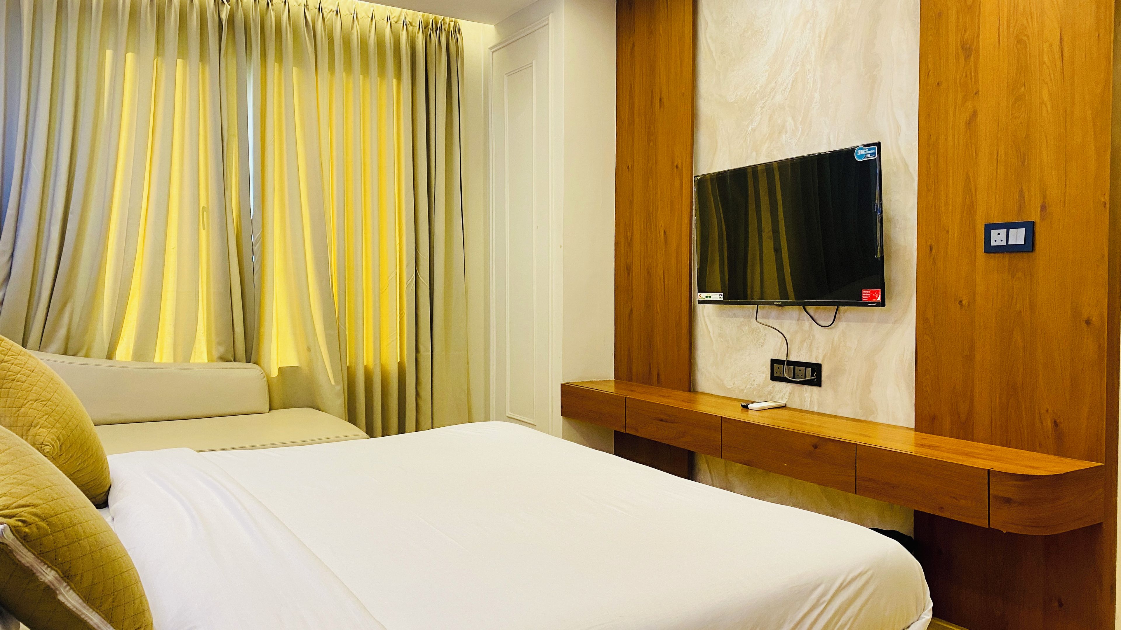 Habitación de hotel con cama, televisor montado en un panel de madera y ventana con cortinas.