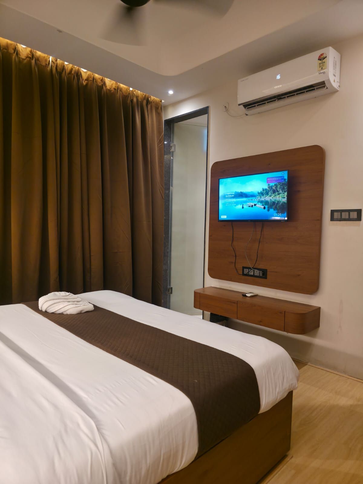 Una habitación de hotel con una cama, un televisor montado en la pared, un equipo de aire acondicionado y un ventanal con cortinas.