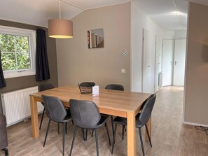 A dining area with a wooden table and four chairs, a hanging light fixture, a window with curtains, and a door leading to another room.