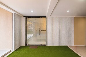 Interior moderno com entrada por porta de vidro, área com grama sintética verde e parede bege com linhas verticais.