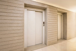 A hallway with two elevator doors, one of which has a digital display. - Hotel O Hanspura Bus Terminal (Ahmedabad)