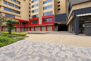 Edifício de vários andares com um pátio, apresentando fachada em vermelho e bege, um caminho pavimentado e uma pequena árvore.