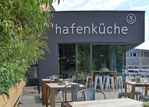 An outdoor seating area with tables and chairs, a building with a sign that reads 'hafenküche,' and a view of the street in the background. - Schiff Grace (Berlin)