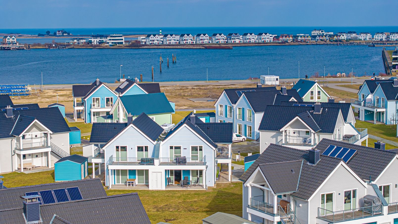 A coastal residential area with houses featuring balconies and solar panels, a pier extending into the water, and a clear view of the sea.