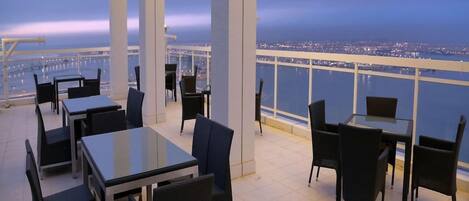 An outdoor terrace with tables and chairs, overlooking a cityscape and water.