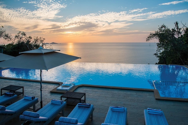 Zona de piscina con sillones y sombrillas, con vista a un cuerpo de agua al atardecer.