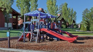 Un parque infantil con un tobogán y estructuras de escalada, rodeado de árboles y un edificio al fondo.