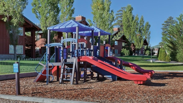 A playground with a slide and climbing structures, surrounded by trees and a building in the background.
