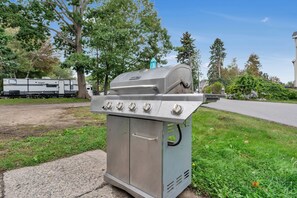 Een draagbare gasgrill met vier branders, een bedieningspaneel en een schoorsteen, geplaatst op een gazon met bomen en een weg op de achtergrond.