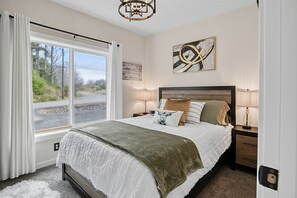 A bedroom with a large window, a bed with multiple pillows, bedside lamps, and a wall art piece. - Sunset View Cottage - Brand new home on the Bluff (Moclips)