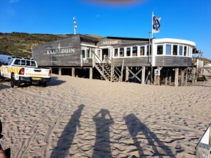 A wooden building with 'KAP DUIN DISHOEK' signage, a white vehicle with 'STRANDWACHT' branding, and a flagpole with a flag.