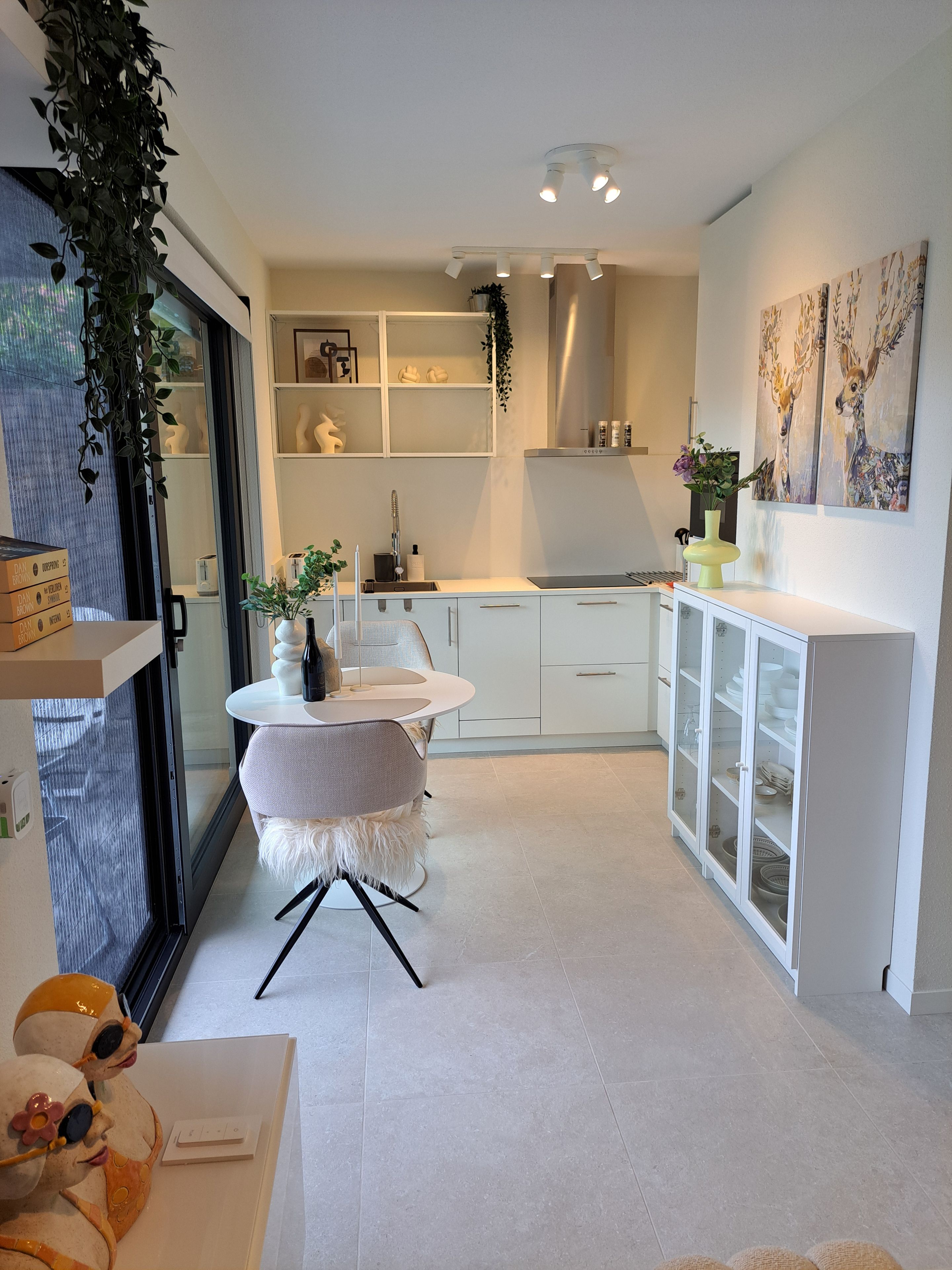 A modern kitchen with white cabinetry, a round table with a fluffy chair, and a glass cabinet with dishes.