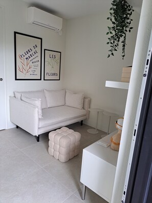 A minimalist living room with a white sofa, a small ottoman, a white side table, and two framed botanical prints on the wall.