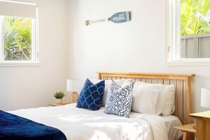 A bedroom with a bed, pillows, a nightstand, a lamp, a wall-mounted sign, and a window with a view of greenery. - "Bluewater" – Pool at home, Beach just beyond (North Avoca)