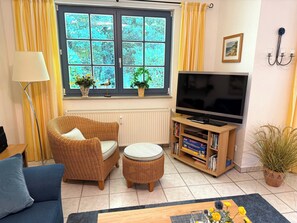 A living room with a television, wicker furniture, a bookshelf, and a window with curtains. - Comfortable vacation rental for up to 4 people, 2 bedrooms, quiet, green setting, west-facing terrace, Wi-Fi, 4-star DTV (Ostseebad Prerow)