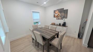 A dining area with a wooden table and chairs, a window with blinds, a coffee machine, and a painting on the wall.