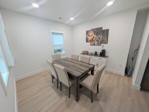 Un coin repas avec une table et des chaises en bois, une fenêtre avec des stores, une machine à café et un tableau accroché au mur.