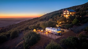 A remote cabin with a large RV parked in front, surrounded by a forested hillside.