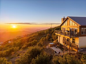 A two-story wooden house with a balcony, surrounded by trees and hills, during sunset.