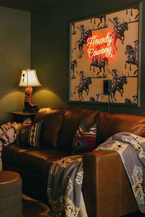 A cozy living room with a leather couch, patterned throw pillows, a lamp, and a neon sign reading "Howdy, Cowboy!" on the wall.