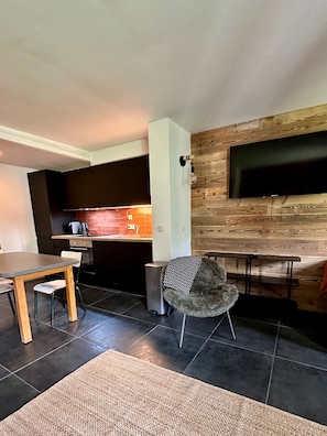 A modern living space with a dining area, a kitchen with a brick backsplash, and a flat-screen TV mounted on a wooden wall. - Plagne 1800, sleeps 6, renovated in 2026, pool, sauna, covered parking. (La Plagne Tarentaise)